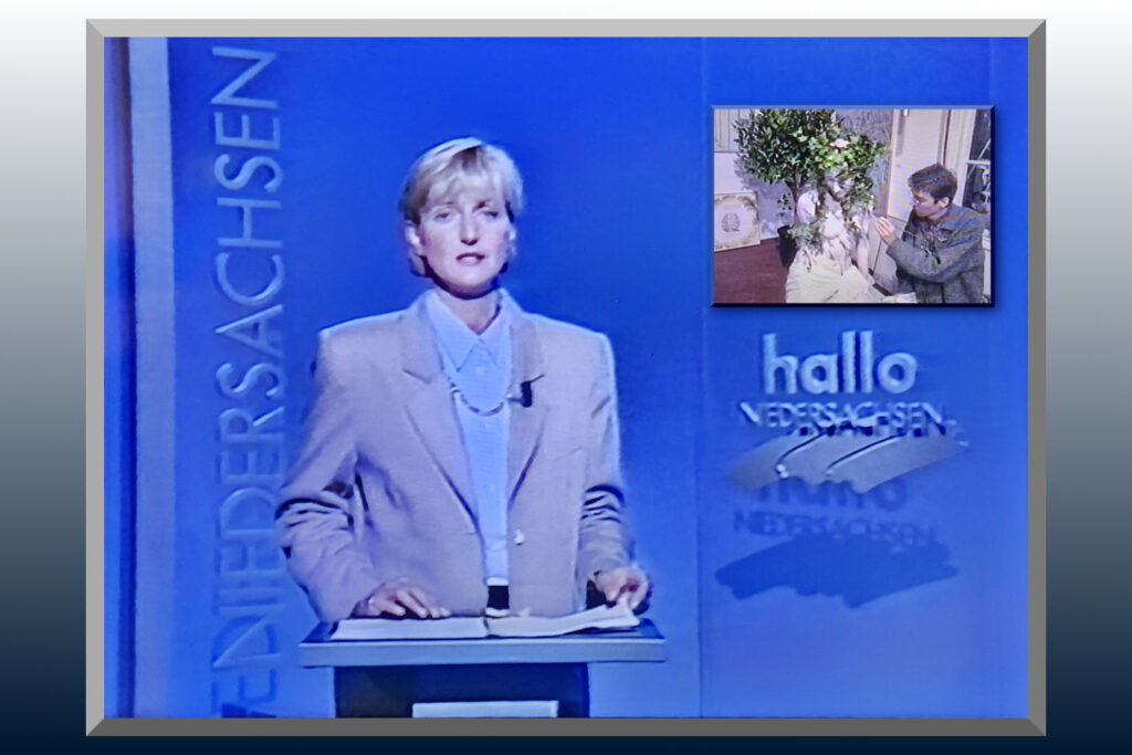 Eine Fernsehmoderatorin steht vor einem Bildschirm, auf dem Künstler Jörg Düsterwald zu sehen ist, während er ein nacktes Fotomodell bemalt.