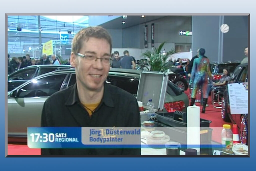 Künstler Jörg Düsterwald wird bei einer Messe von einem Fernsehteam begleitet und interviewt.