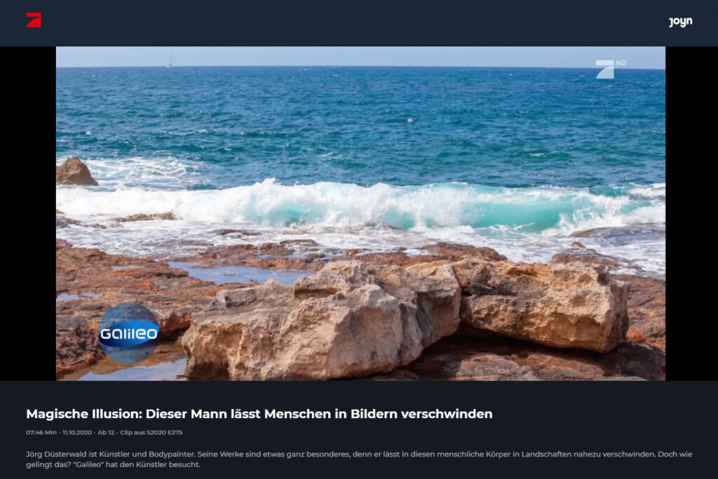 Ein wie ein Stein bemaltes Fotomodell liegt vor der Brandung an einem Strand auf Felsen.