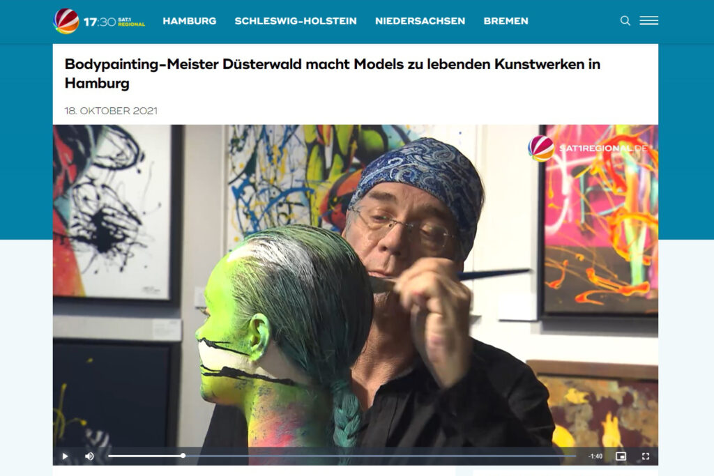 Körperkünstler Jörg Düsterwald bemalt in einer Galerie mit Bildern den Kopf eines Fotomodells.