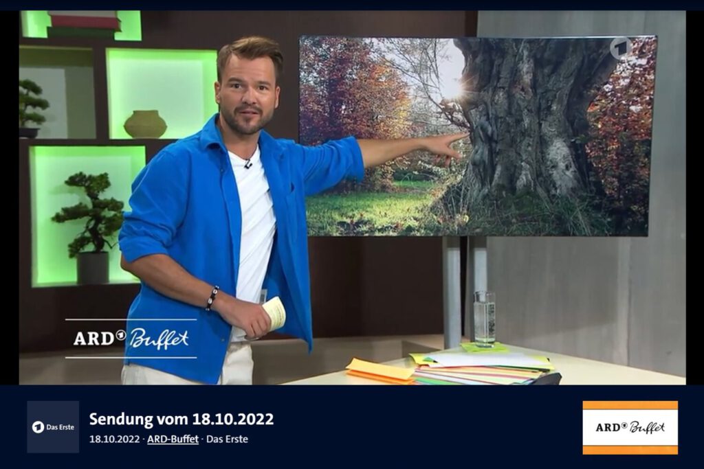 Der Moderator einer Fernsehsendung zeigt auf einen Bildschirm im Hintergrund.