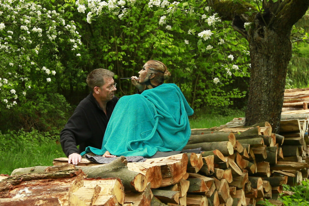 Ein Fotomodell sitzt mit einem Bademantel bedeckt auf einem Holzstapel. Die Frau wird für das Kunstprojekt Naturart von Künstler Jörg Düsterwald bemalt.