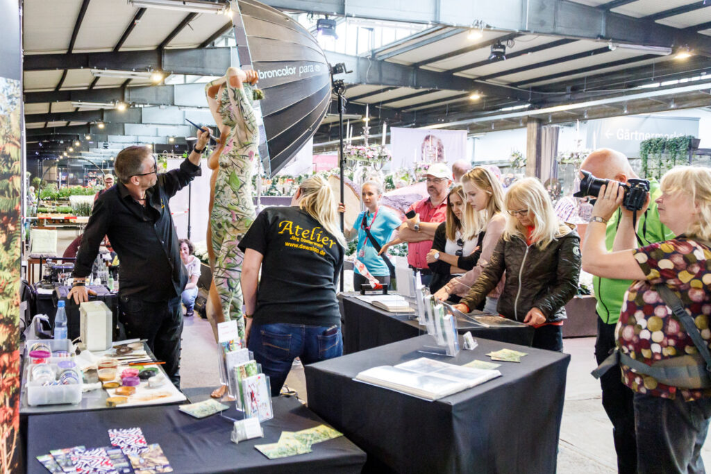 Künstler Jörg Düsterwald bemalt zusammen mit einer Assistentin ein haltnacktes Fotomodell mit einem Blumenmuster. Sind befinden sich auf einer Messeausstellung, Publikum schaut den Dreien zu.