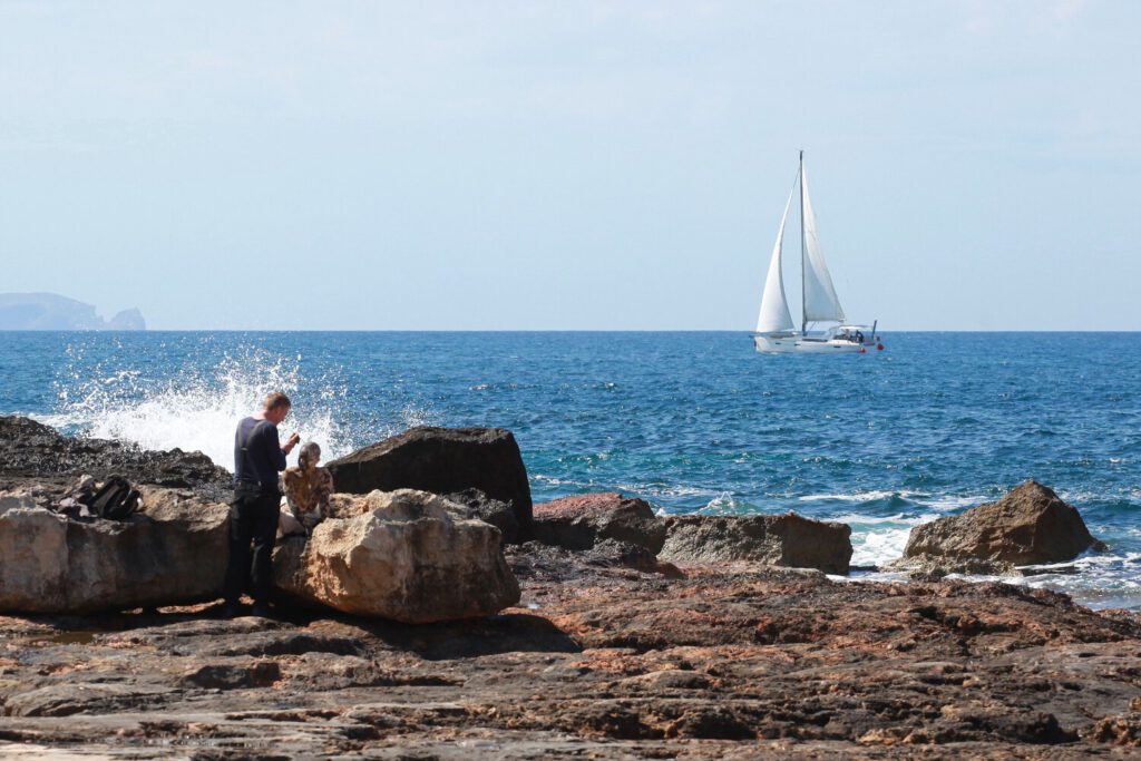 Ein nacktes Fotomodell wird für das Kunstprojekt Natureart von Künstler Jörg Düsterwald vollständig so mit Farbe bemalt, dass die Person mit der Kulisse verschmilzt. Sie sind auf der Insel Mallorca. Die Frau sitzt auf einem Felsen am Meer, der Künstler steht vor ihr. Wellengischt spritzt hoch, im Hintergrund segelt ein Boot vorbei.