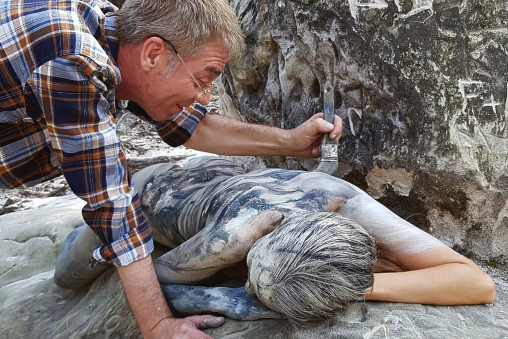 Ein nacktes Fotomodell wird für das Kunstprojekt Natureart von Künstler Jörg Düsterwald vollständig so mit Farbe bemalt, dass die Person mit der Kulisse verschmilzt. Sie liegt an einem Felsen, der Künstler hat einen Pinsel in der Hand und beugt sich über sie.