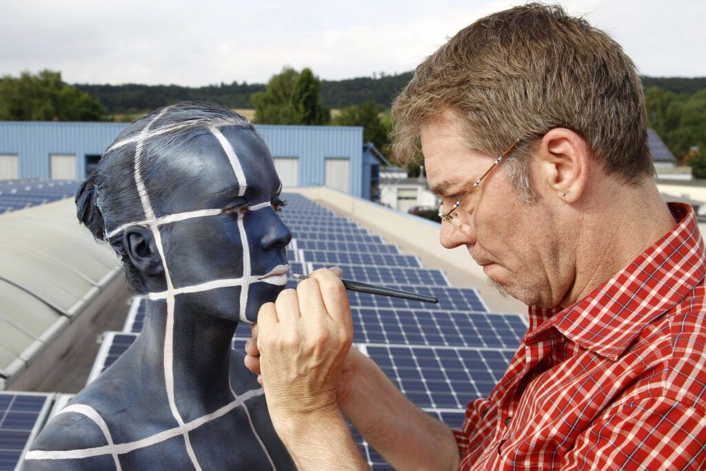 Künstler Jörg Düsterwald und ein Fotomodell befinden sich auf einem Gebäudedach mit einer Solaranlage. Der Künstler bemalt die Frau mit dem Muster der Photovoltaikanlage.