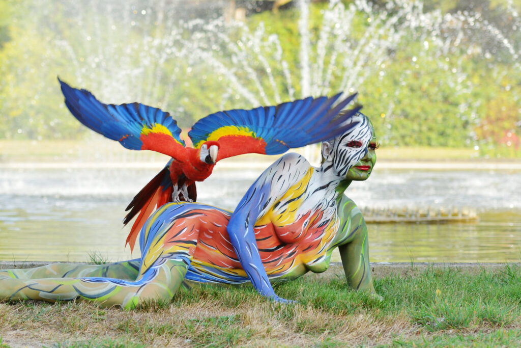 Ein Fotomodell wurde für das Kunstprojekt ANIMAL ART von Künstler Jörg Düsterwald vollständig bunt bemalt. Die Frau posiert mit Papageien. Ein Vogel fliegt davon.