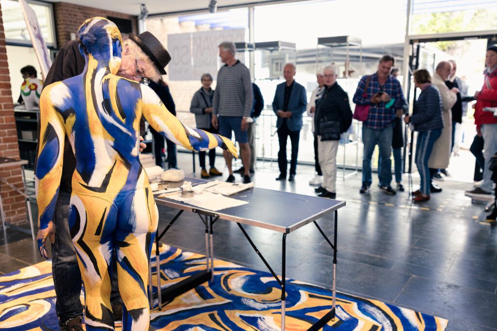 Künstler Jörg Düsterwald bemalt bei einem Kunstevent eine nackte Frau mit blaugelber Farbe. Publikum steht drum herum und beobachtet die Aktion.