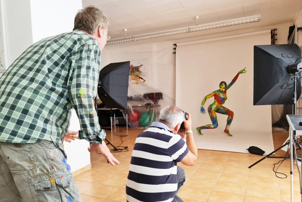Künstler Jörg Düsterwald ist mit einem Bodypaintingmodell und einem Fotografen bei einer Foto-Performance in seinem Atelier. Die Frau posiert vor einer Kulisse, der Künstler gibt Anweisungen, während der Fotograf mit Scheinwerfen und Kamera Fotos macht.