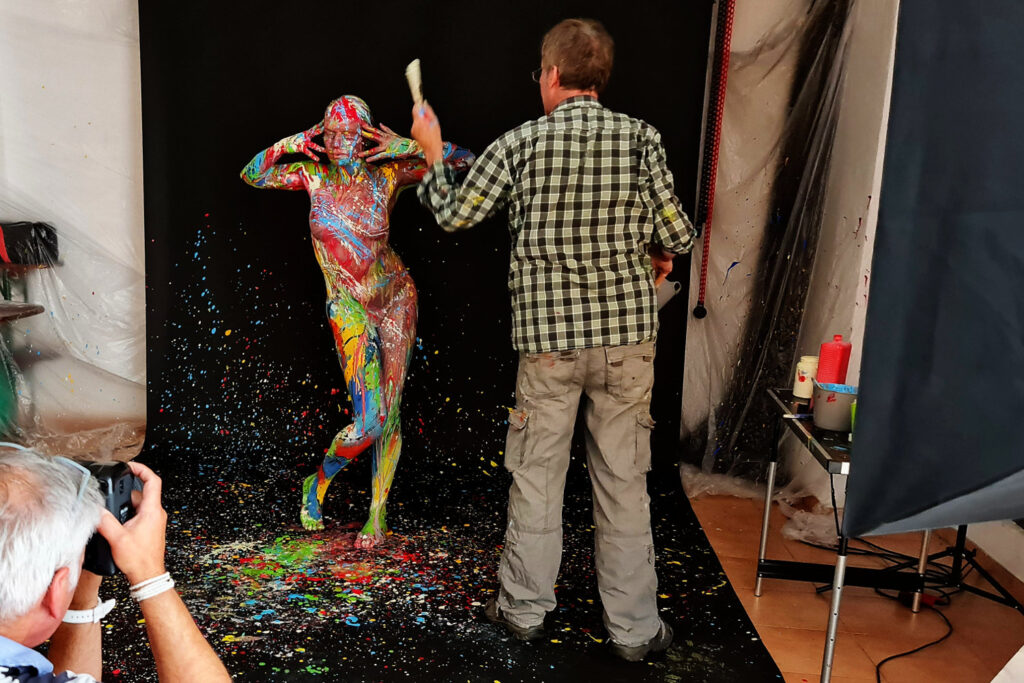 Künstler Jörg Düsterwald ist mit einem bunt bemalten, nackten Fotomodell in seinem Atelier. Die Frau steht vor einer Fotokulisse und der Künstler vor ihr. Er hat einen Pinsel in der Hand und bekleckert die Frau mit Farbe. Ein Fotograf fotografiert die Szene.