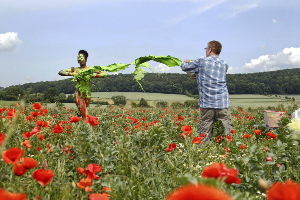 Künstler Jörg Düsterwald ist mit einer bunten bemalten nackten Frau in einem Mohnfeld. Für ein Fotomotiv bewirft er die Person mit grüner flüssiger Farbe.