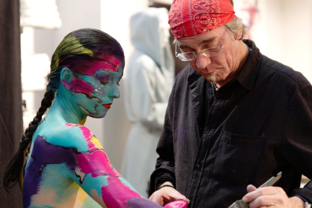 Künstler Jörg Düsterwald trägt ein rotes Kopftuch und bemalt ein Modell mit bunter Farbe.