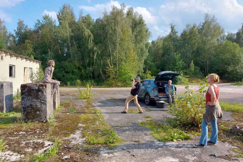 Künstler Jörg Düsterwald hat ein nacktes Fotomodell vollständig mit Farbe bemalt. Sie sitzt in einem Industriegelände auf einem Stein und ein Fotograf macht Fotos von Ihr. Der Künstler und eine Assistentin schauen zu.