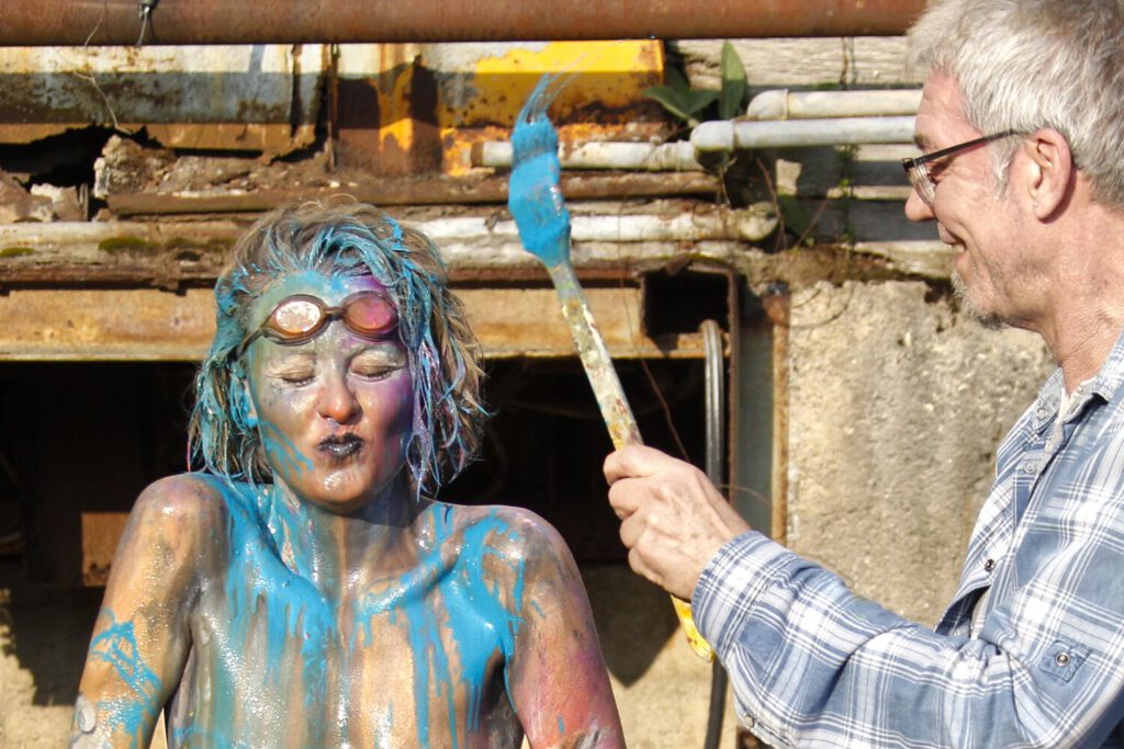 Künstler Jörg Düsterwald ist mit einer bunten bemalten nackten Frau an einem alten Industriegebäude. Er hat einen Pinsel mit blauer Farbe in der Hand und möchte die Frau bespritzen. Sie kneift die Augen zu und spitzt schmunzelnd die Lippen
