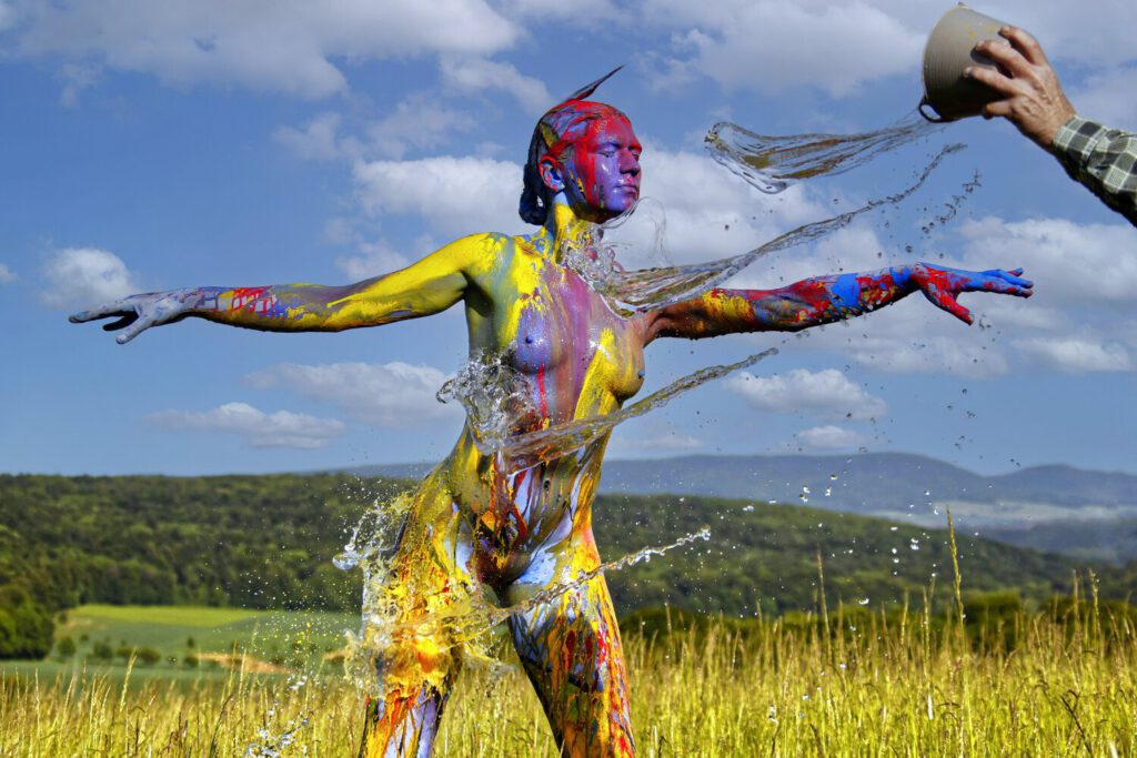 Künstler Jörg Düsterwald hat für das Kunstprojekt Splashart ein nacktes Fotomodell vollständig mit bunter Farbe bemalt. Die Frau steht mit ausgestreckten Armen in einer Wiese, der Künstler kippt aus einer kleinen Schale Wasser über sie.