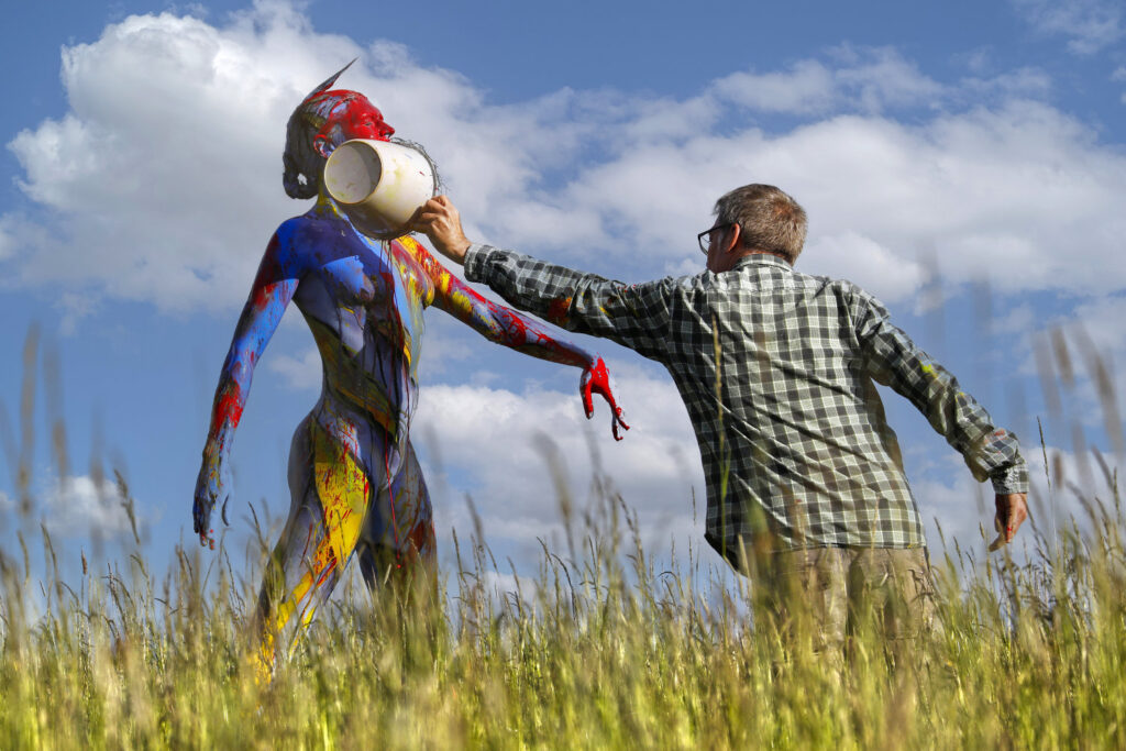 Künstler Jörg Düsterwald hat für das Kunstprojekt Splashart ein nacktes Fotomodell vollständig mit bunter Farbe bemalt. Die Frau steht mit ausgestreckten Armen in einer Wiese, der Künstler kippt aus einem kleinen Eimer Farbe über ihre Brust.