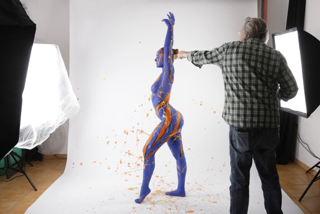 Eine nackte, von Künstler Jörg Düsterwald mit bunter Farbe versehene Frau steht seitlich, sich reckend in einem Fotostudio. Der Künstler steht seitlich hinter ihr, hält einen Pinsel mit orangener Farbe in der Hand und besprenkelt sie damit.