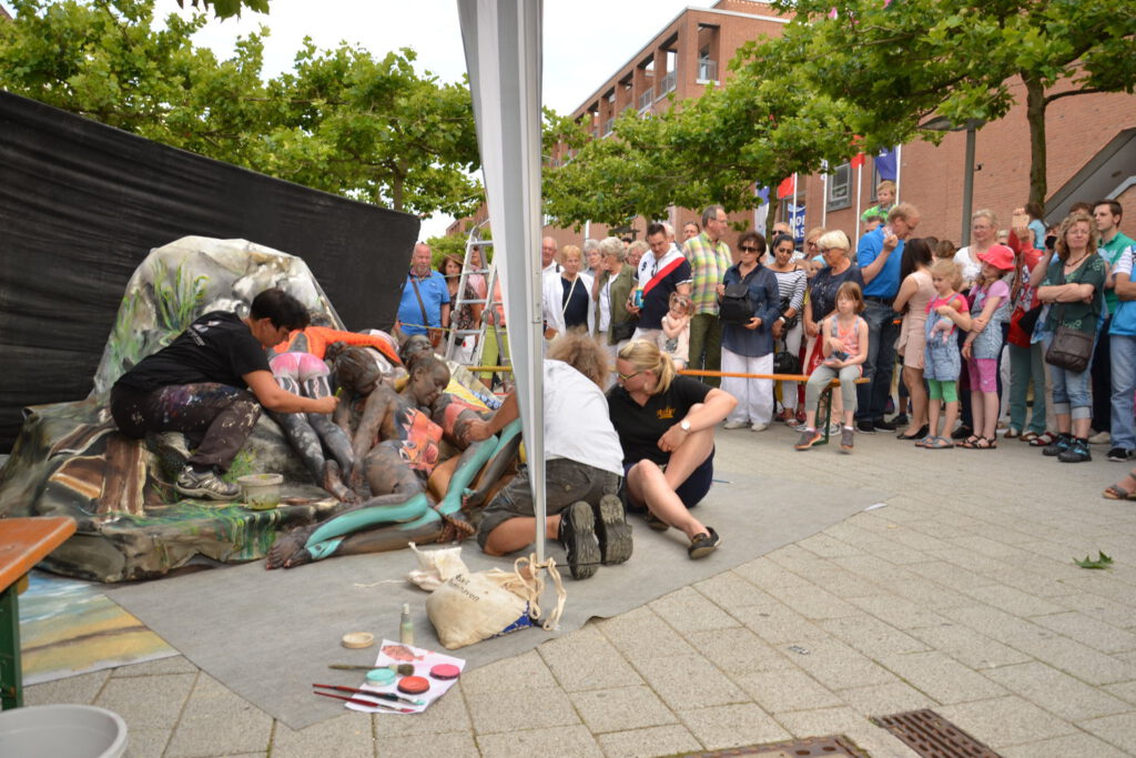 Bei einem Straßenkunst-Fest inszeniert Künstler Jörg Düsterwald zusammen mit Kolleginnen und halbnackten Modellen eine Kunstperformance. Die Modelle liegen auf einem Podest, die Künstler bemalen die Frauen. Viel Publikum steht herum und beobachtet das Geschehen.