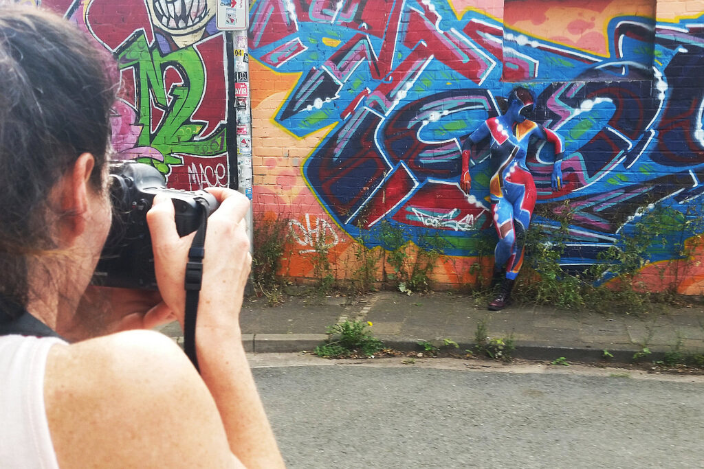 Künstler Jörg Düsterwald hat eine nackte Frau vollständig und bunt mit Farbe bemalt. Sie steht vor einer Graffitiwand, eine Fotografin fotografiert die Person.