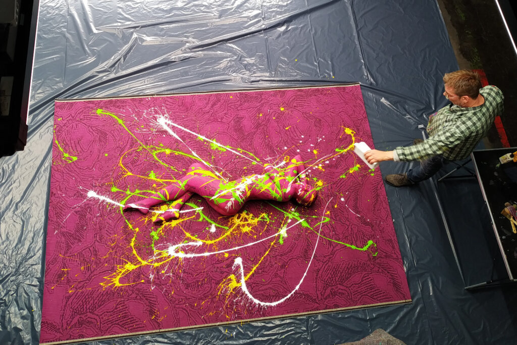 Ein magentafarbener Teppich liegt auf dem Boden. Für ein Teppichprojekt hat Künstler Jörg Düsterwald ein nacktes Fotomodell vollständig im gleichen Farbton bemalt. Die Frau liegt seitlich auf dem Teppich, der Künstler steht seitlich daneben und besprenkelt sie mit bunten Farben.