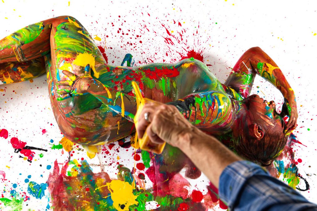 Eine nackte, von Künstler Jörg Düsterwald mit bunter Farbe versehene Frau liegt seitlich auf dem Boden. Der Künstler hält eine Flasche mit gelber Farbe über ihr in der Hand und besprenkelt sie damit.