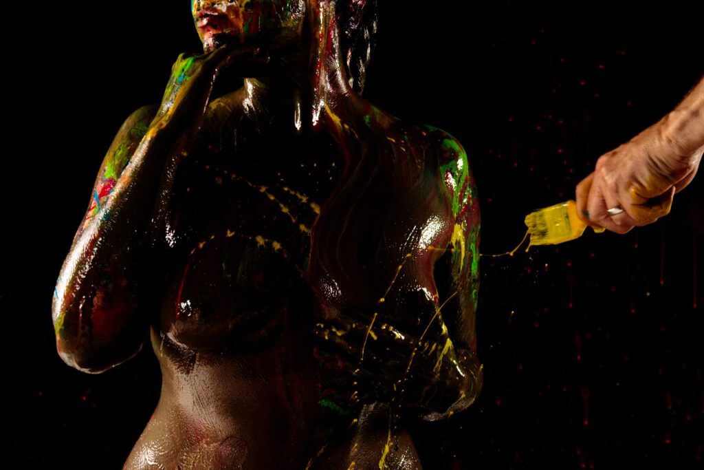 Künstler Jörg Düsterwald hat den Oberkörper und das Gesicht eines nackten Fotomodells mit dunkler Farbe versehen. Er hält einen Pinsel mit gelber Farbe in der Hand und bekleckert damit die Person.