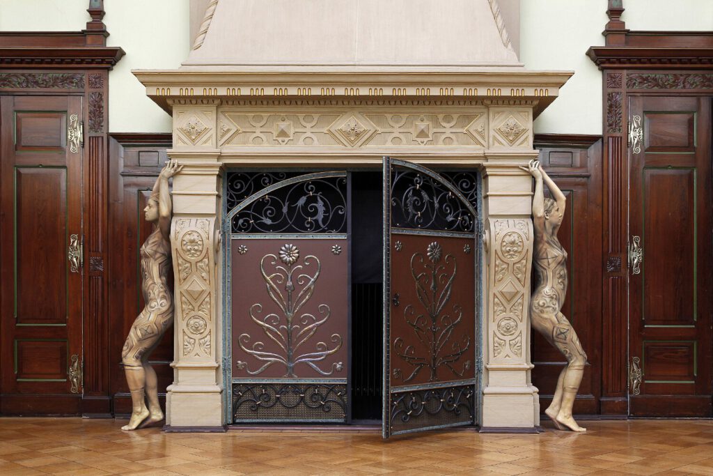 Für das Kunstprojekt DOOR ART ließen sich zwei nackte Fotomodelle von Künstler Jörg Düsterwald vollständig so mit Farbe bemalen, dass sie genau zur Umgebung von Türen und Toren passen. Sie stehen wie Steinskulpturen links und rechts neben einem großen Kamin.