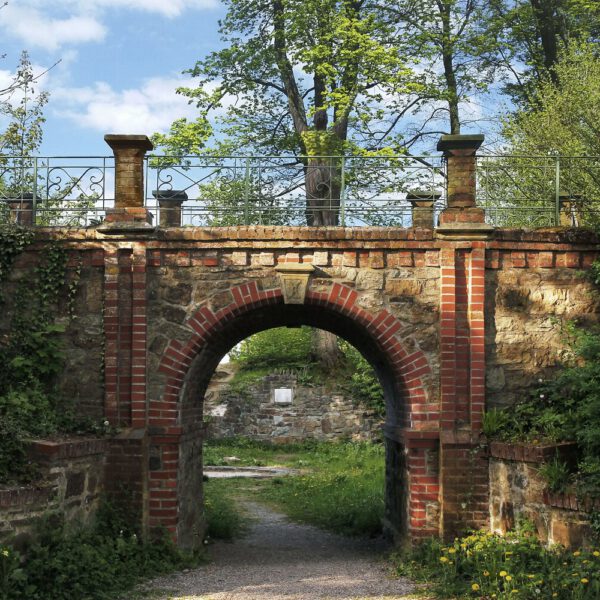 Für das Kunstprojekt DOOR ART ließ sich ein nacktes Fotomodell von Künstler Jörg Düsterwald vollständig so mit Farbe bemalen, dass es genau zur Umgebung von Türen und Toren passt. Die Frau steht über einem Steintorbogen vor einem Baum und ist kaum zu sehen.