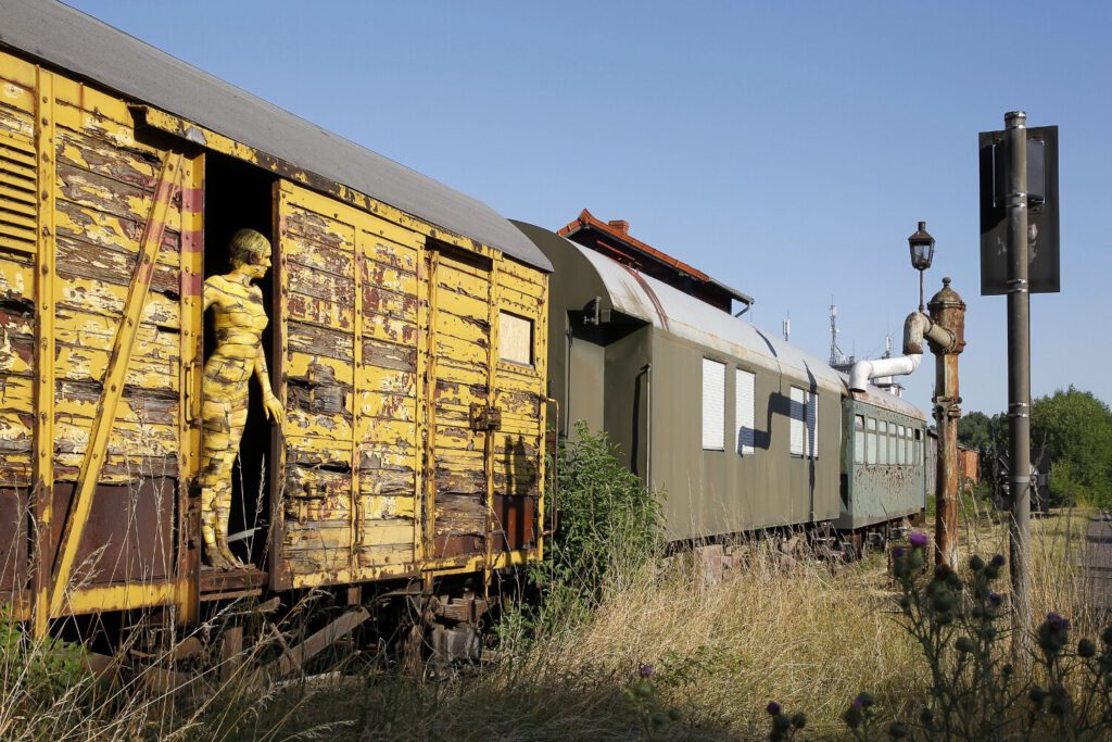 Für das Kunstprojekt DOOR ART ließ sich ein nacktes Fotomodell von Künstler Jörg Düsterwald vollständig so mit Farbe bemalen, dass es genau zur Umgebung von Türen und Toren passt. Die Frau steht in der offenen Tür eines alten, gelben Güterwaggons auf einem verlassenen Bahnhof.