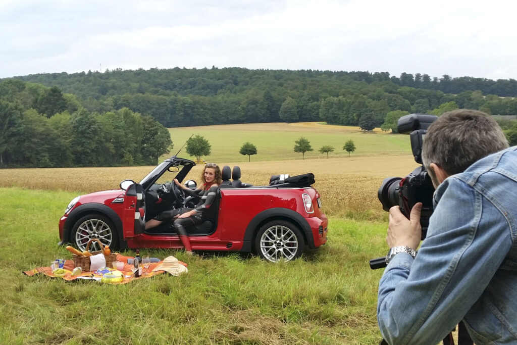 Ein Auto mit offener Fahrertür steht in einer Wiese, davor ist eine Picknickdecke ausgebreitet. Im Auto sitzt ein nacktes Fotomodell und wurde von Künstler Jörg Düsterwald so mit Farbe bemalt wie das Auto von innen. Ein Fotograf fotografiert die Szene.