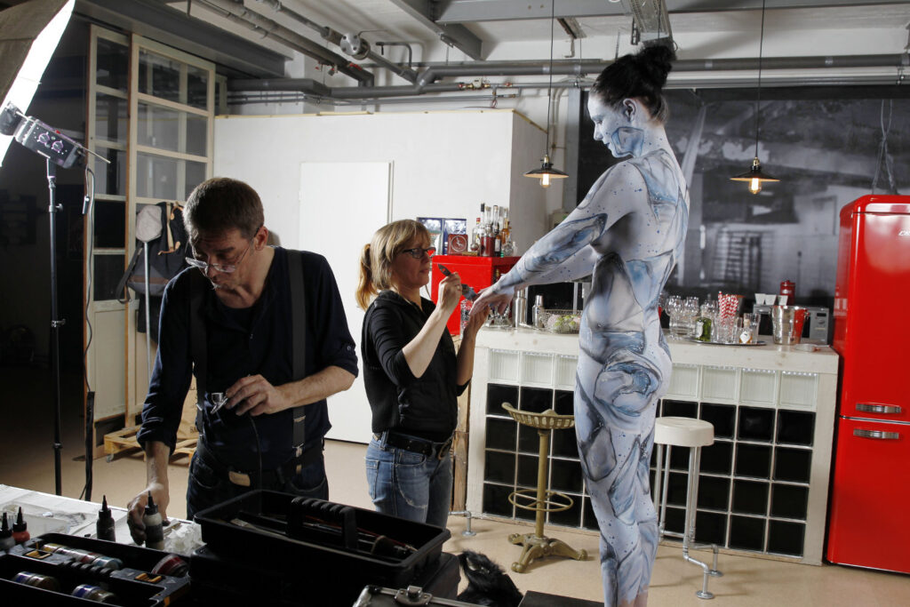 Ein nacktes Fotomodell steht in der Nähe eines großen roten Kühlschranks. Künstler Jörg Düsterwald hat die Frau vollständig mit Farbe bemalt, das Motiv sind Eiswürfel. Eine Assistentin hilft dabei.