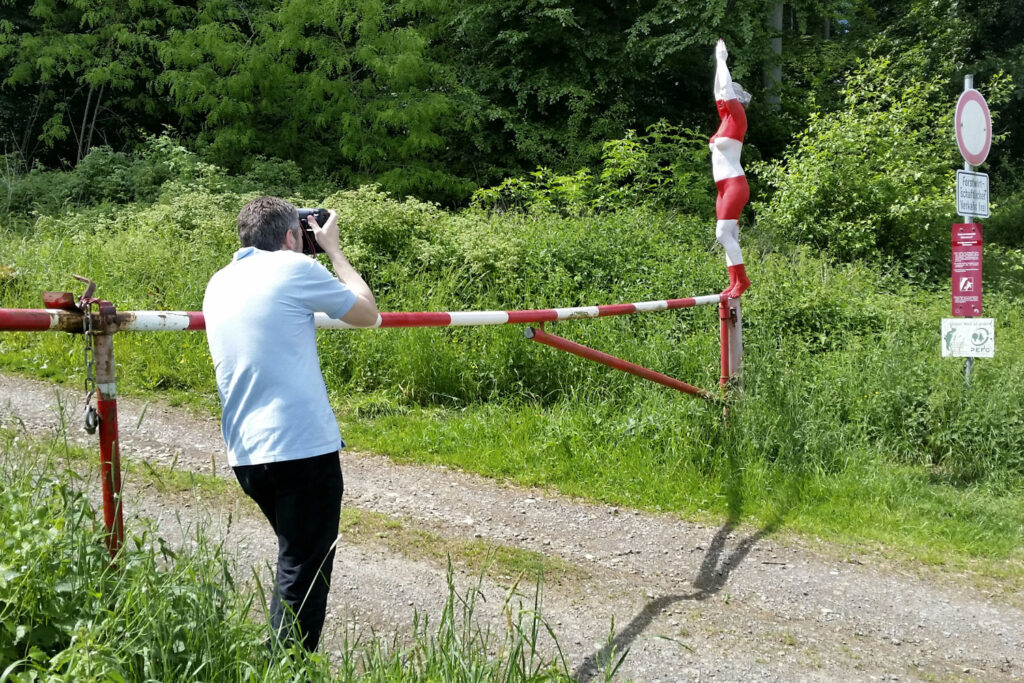 Ein nacktes Fotomodell wurde von Künstler Jörg Düsterwald vollständig so mit Farbe bemalt, dass die Person mit der Kulisse verschmilzt. Sie steht in einem bunten Blumenfeld und wird von einem Fotografen fotografiert.