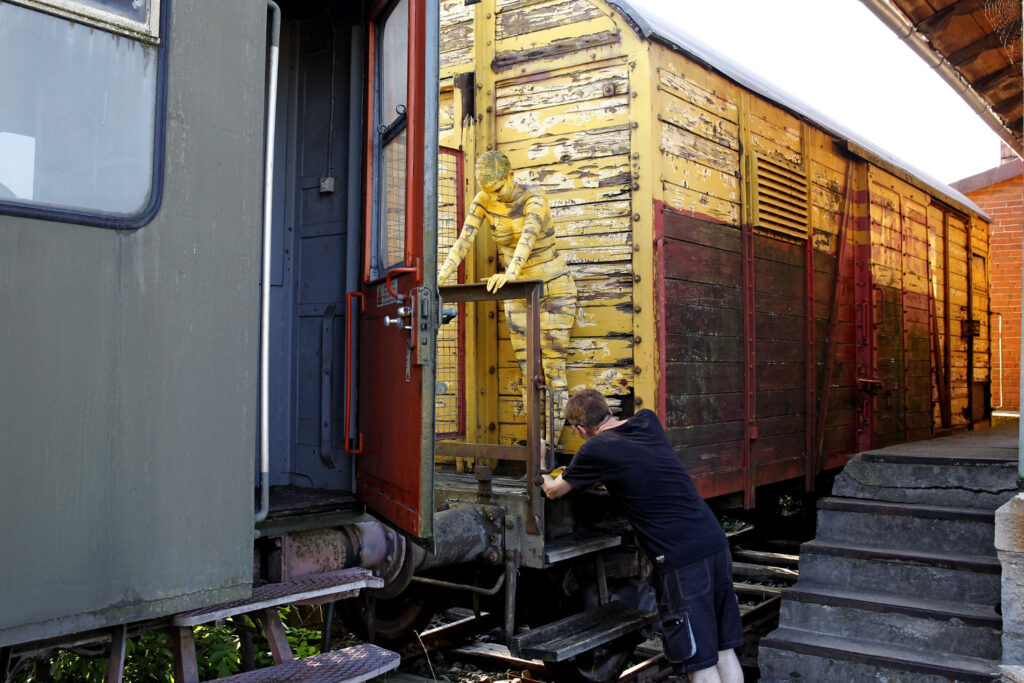 Ein nacktes Fotomodell steht an einem schäbbigen, gelben Güterwaggon. Künstler Jörg Düsterwald bemalt die Frau vollständig so mit Farbe, dass die Person mit der Kulisse verschmilzt.