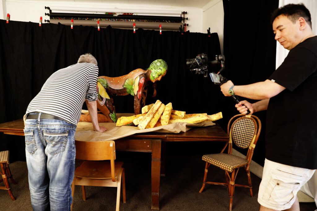 Künstler Jörg Düsterwald hat mit einem nackten Fotomodell einen Hamburger kreiert. Zur Dekoration liegen überdimensionale PommesFrites auf dem Tisch. Ein Kameramann filmt die Szene.
