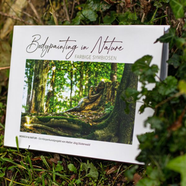 Künstler Jörg Düsterwald hat von dem Körperkunstprojekt Nature Art einen Fotobildband publiziert. Das Buch liegt im Gras an einem Baumstamm.