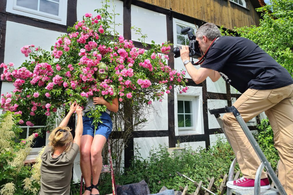Für das Kunstprojekt Fowerart hat Künstler Jörg Düsterwald ein Fotomodell im Gesicht mit einem rosa Rosenmuster bemalt. Die Frau steht vor einem Rosenbusch, eine Assistentin hält Zweige und ein Fotograf fotografiert von einer Leiter aus die Szene.