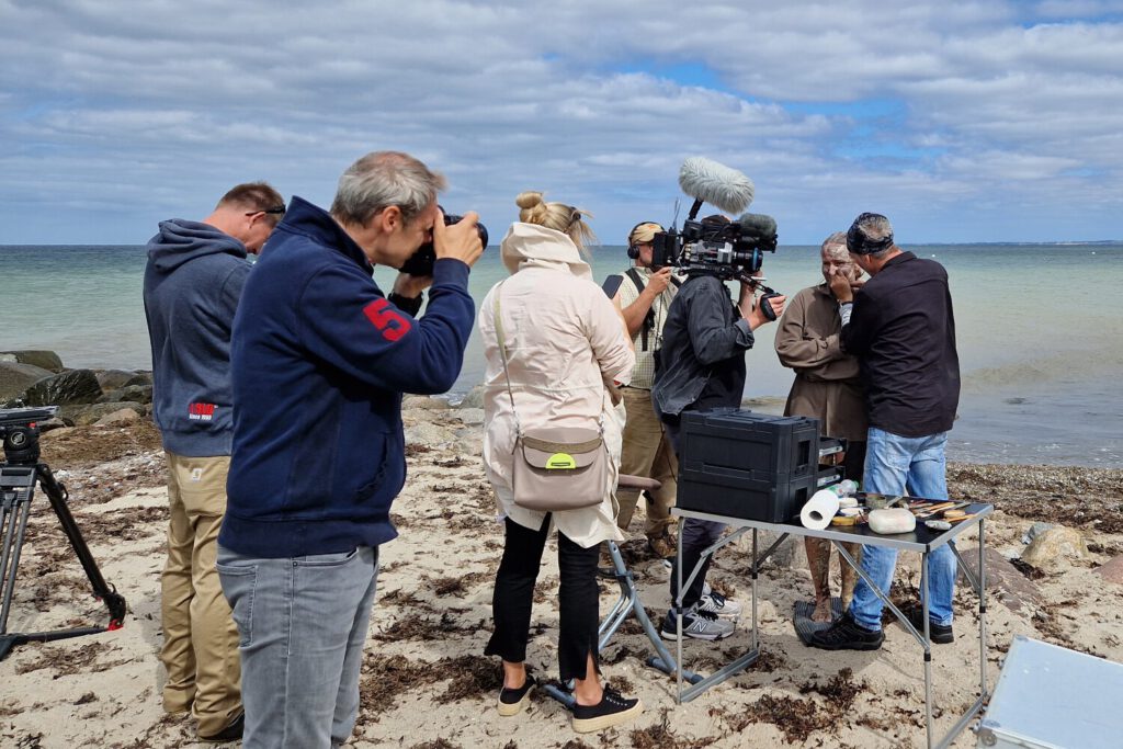 Ein Fotomodell wird von Künstler Jörg Düsterwald im Gesicht mit Farbe bemalt. Sie stehen an einem Strand, ein Kamerateam eines Fernsehsenders filmt das Geschehen. Ein Fotograf steht daneben und fotografiert.