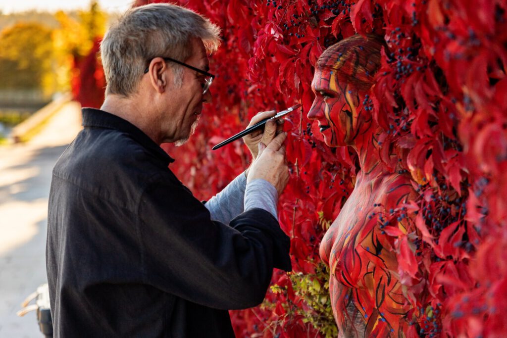 Ein nacktes Fotomodell steht zwischen den roten Blättern einer Weinrankenwand. Künstler Jörg Düsterwald hält einen Pinsel mit roter Farbe in der Hand und bemalt die Frau vollständig so mit Farbe, dass sie mit der Kulisse verschmilzt.