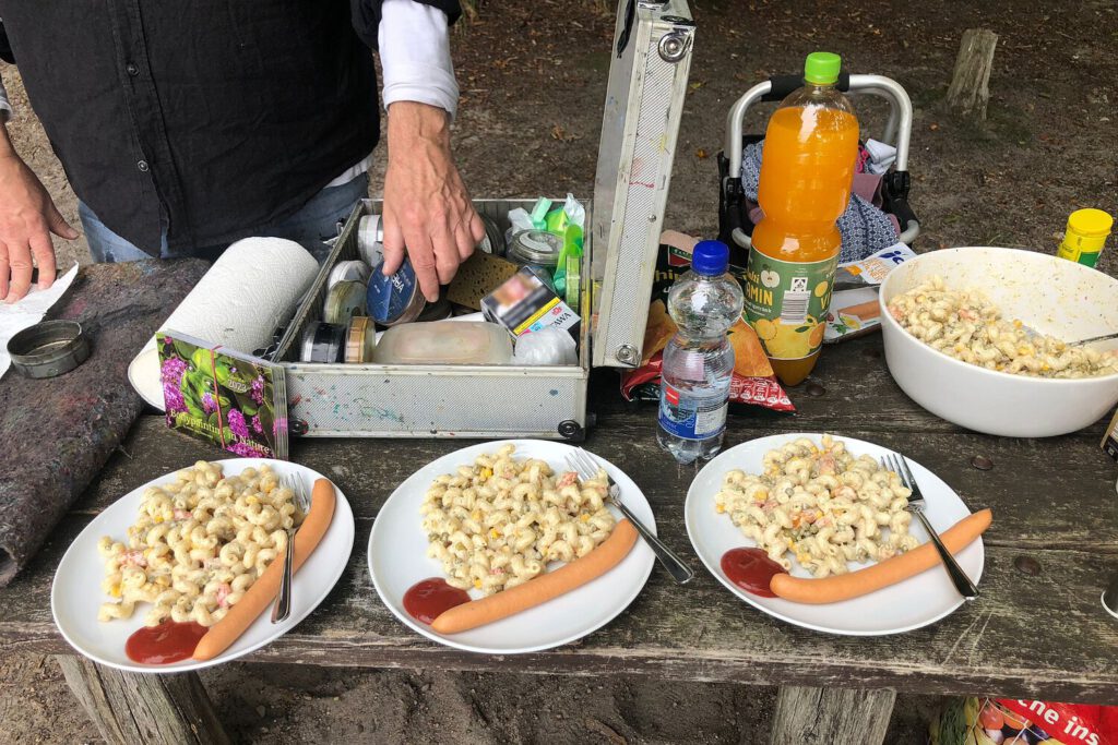 Auf einem Waldtisch stehen Picknickteller mit Kartoffelsalat, Würstchen und Ketchup