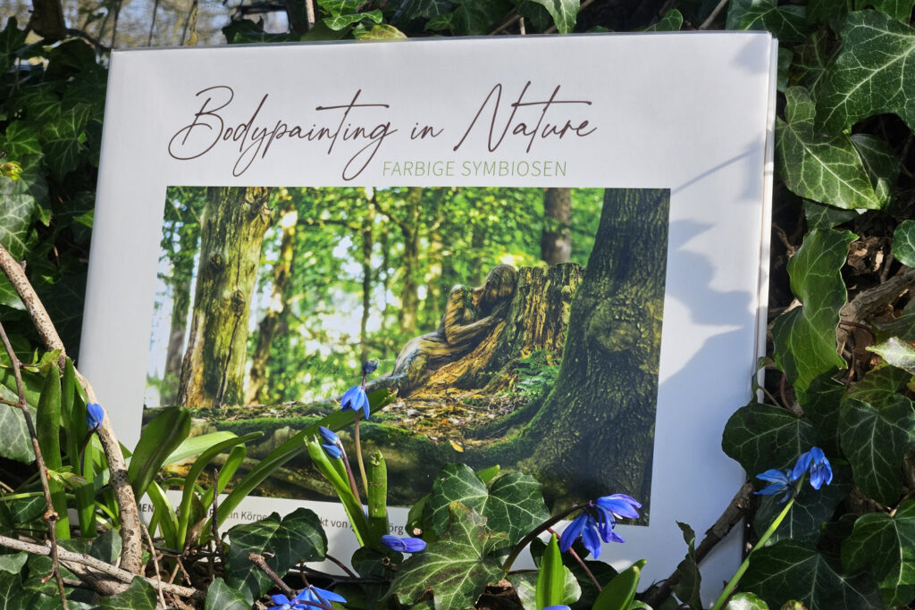Künstler Jörg Düsterwald hat von dem Körperkunstprojekt Nature Art einen Fotobildband publiziert. Das Buch liegt zwischen Frühlingsblumen an einem Baumstamm.