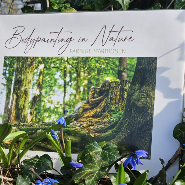 Künstler Jörg Düsterwald hat von dem Körperkunstprojekt Nature Art einen Fotobildband publiziert. Das Buch liegt zwischen Frühlingsblumen an einem Baumstamm.