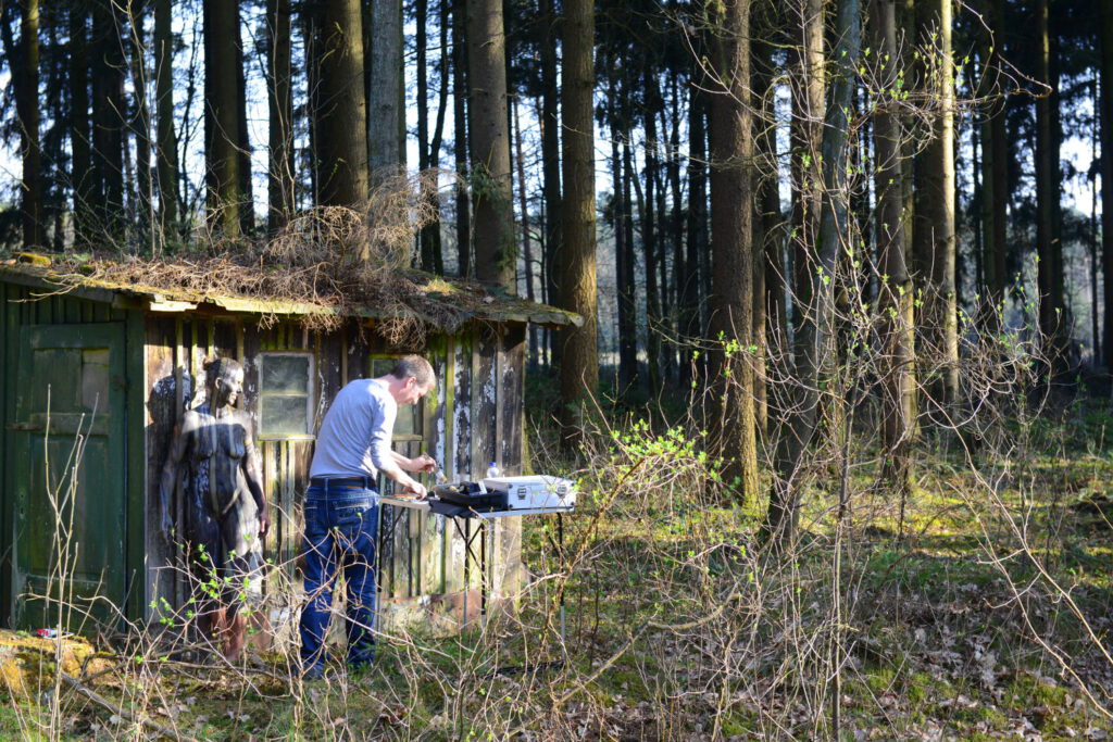 Ein nacktes Fotomodell wird für das Kunstprojekt Natureart von Künstler Jörg Düsterwald vollständig so mit Farbe bemalt, dass die Person mit der Kulisse verschmilzt. Die Frau steht im Wald an einer Holzhütte, der Künstler steht mit einem Tisch und einem Pinsel in der Hand bei ihr.