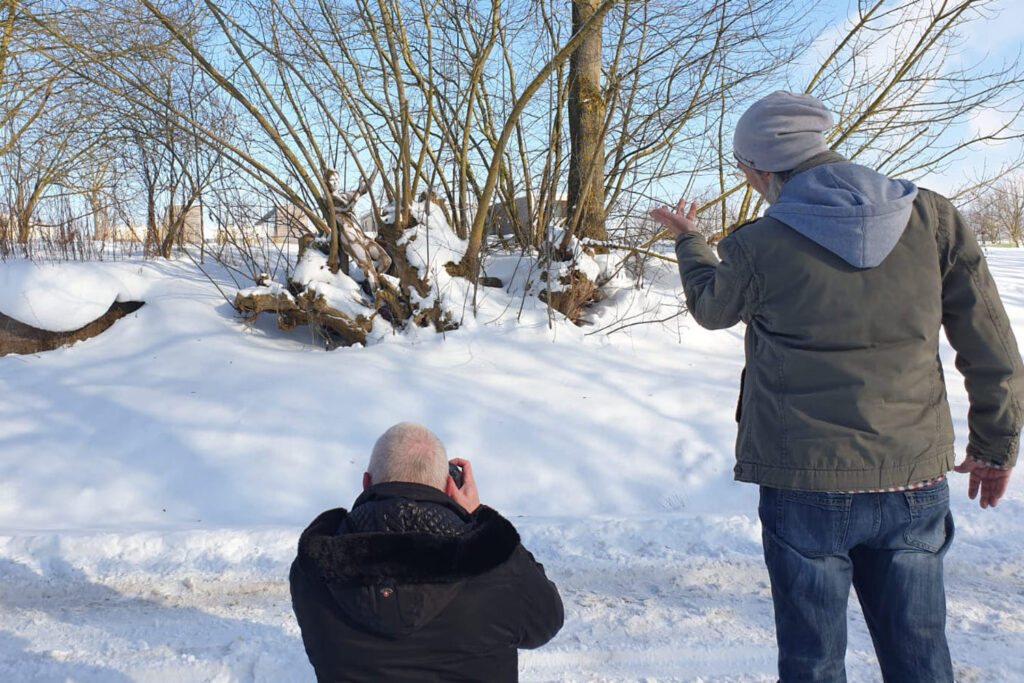 Künstler Jörg Düsterald hat ein nacktes Fotomodell vollständig mit Farbe bemalt, sodass die Frau mit der Kulisse verschmilzt. Sie steht in einer Schneelandschaft zwischen Bäumen, ein Fotograf fotografiert knieend die Szene. Der Künstler steht daneben und gibt Anweisungen.