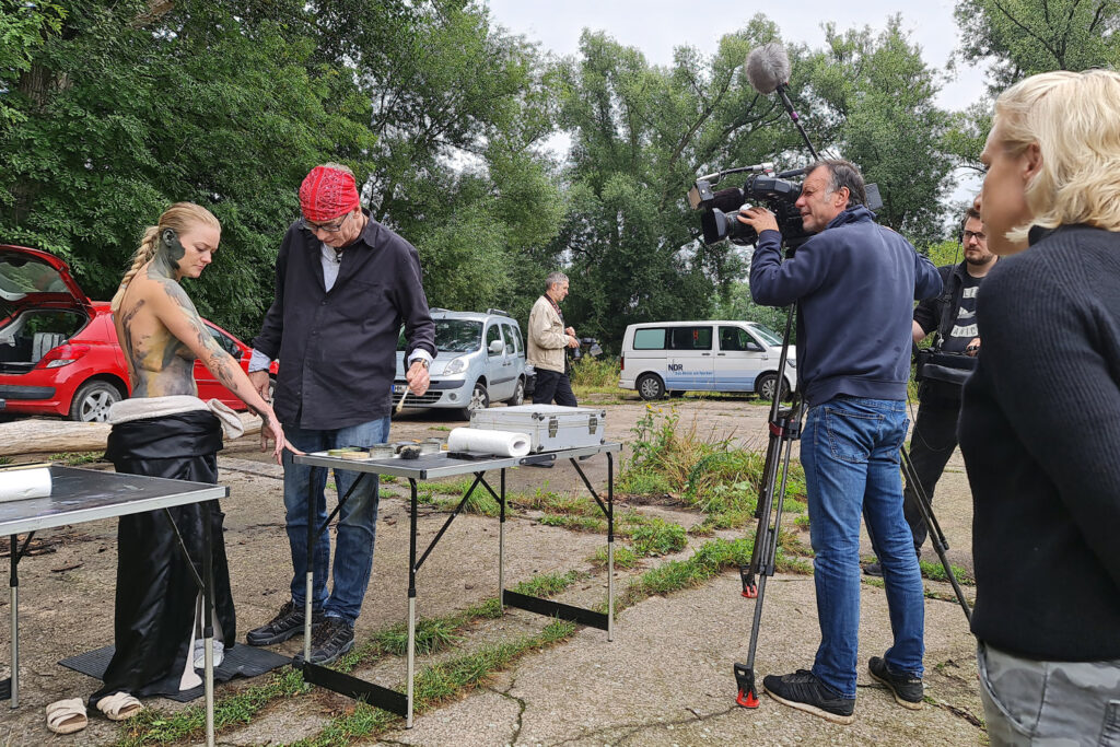 Ein nacktes Fotomodell wird von Künstler Jörg Düsterwald mit Farbe bemalt. Sie stehen an einem Waldrand und ein Kamerateam eines Fernsehsenders filmt das Geschehen.