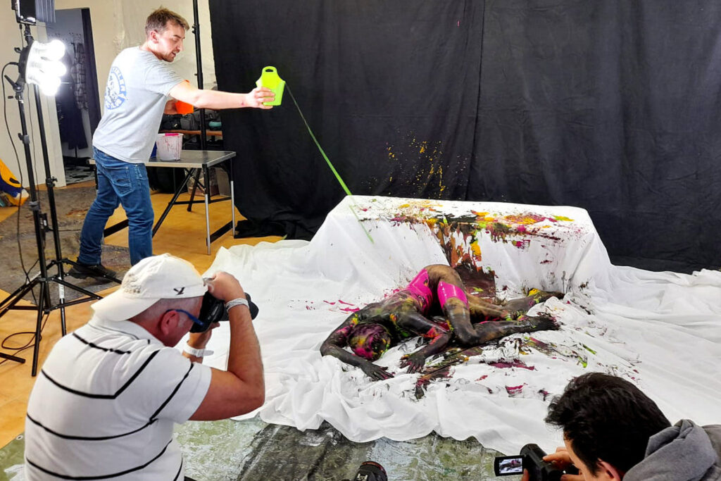 Eine von Künstler Jörg Düsterwald bunte bemalte nackte Frau liegt in einem Fotostudio auf einem weißen Tuch. Ein Assistent übergießt die Person mit grüner flüssiger Farbe, ein Fotograf fotografiert die Aktion.
