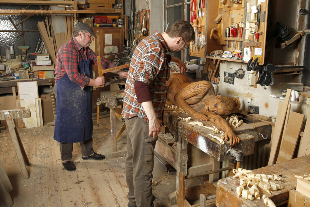 In einer alten Tischlerwerkstatt wurde für das Projekt Workingart ein nacktes Fotomodell von Künstler Jörg Düsterwald vollständig mit einem Holzmuster bemalt. Die Frau liegt auf einer Hobelbank, der Künstler arrangiert letzte Details. Im Hintergrund steht ein Tischler und sägt an einem Brett.