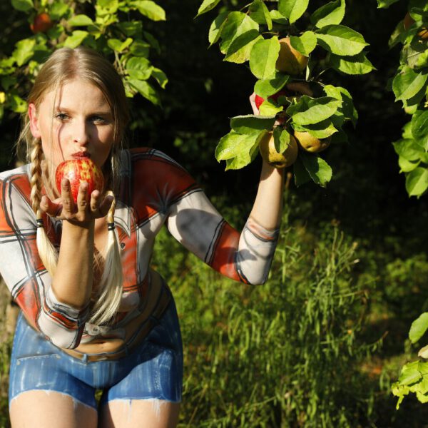 Im Rahmen des Kunstprojektes AGRAR ART hat Bodypaint-Künstler Jörg Düsterwald ein nacktes Fotomodell mit Körperfarbe so bemalt, als hätte die Frau eine kurze blaue Hose und ein rotweiß-kariertes Hemd an. Sie befindet sich in einer Apfelbaumplantage und erntet die Äpfel.