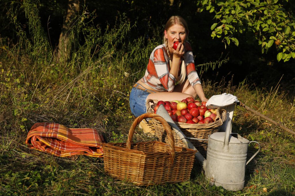 Im Rahmen des Kunstprojektes AGRAR ART hat Bodypaint-Künstler Jörg Düsterwald ein nacktes Fotomodell mit Körperfarbe so bemalt, als hätte die Frau eine kurze blaue Hose und ein rotweiß-kariertes Hemd an. Sie befindet sich in einer Apfelbaumplantage und erntet die Äpfel.