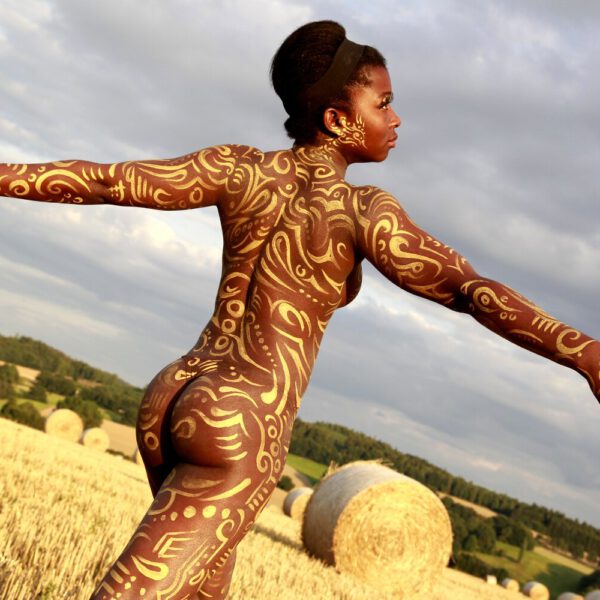 Für das Kunstprojekt AGRAR ART hat Bodypaint-Künstler Jörg Düsterwald mit Körperfarbe ein dunkelhäutiges Fotomodell mit goldfarbenen Ornamenten bemalt. Die Frau posiert in der Abendsonne bei Strohballen auf einem Strohfeld.