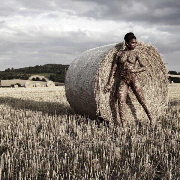Für das Kunstprojekt AGRAR ART hat Bodypaint-Künstler Jörg Düsterwald mit Körperfarbe ein dunkelhäutiges Fotomodell mit goldfarbenen Ornamenten bemalt. Die Frau posiert in der Abendsonne bei Strohballen auf einem Strohfeld.